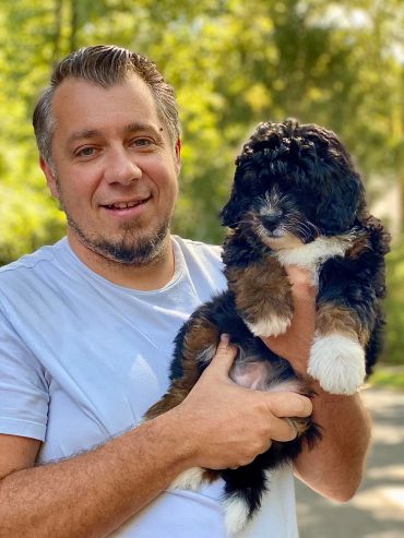 Man holding a fluffy black, brown, and white puppy outdoors.