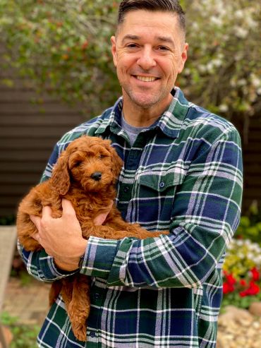 Man smiling while holding a fluffy brown puppy outdoors.
