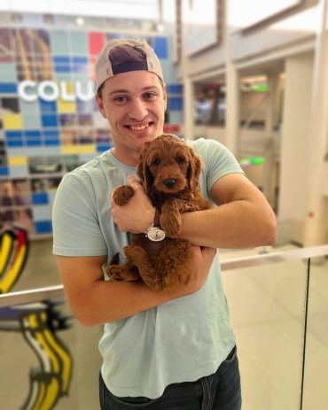 Young man holding a fluffy brown puppy indoors, smiling warmly.