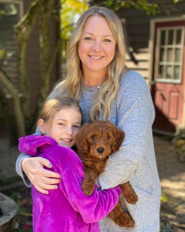 Woman and girl hugging a brown puppy outdoors with smiles.