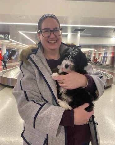 A smiling woman in a gray coat holding a black and white puppy at an airport baggage claim.