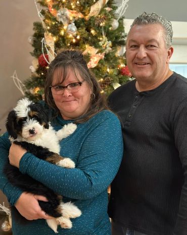 A couple smiling and holding a fluffy puppy by a decorated Christmas tree.