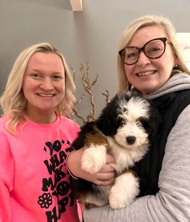 Two women smiling, one holding a fluffy black, white, and brown puppy indoors.