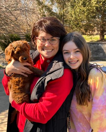 Two smiling women outdoors with a brown puppy on a sunny day.