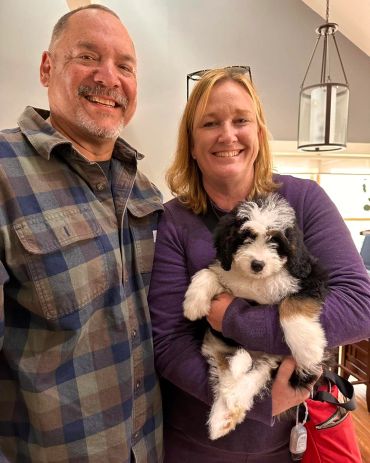 A happy couple holding a fluffy black, white, and brown puppy indoors.