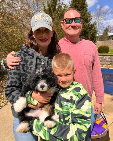 Family of three posing happily outdoors with their playful black and white puppy.