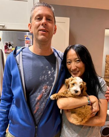A couple smiling, holding a fluffy puppy indoors.
