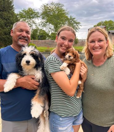 Family smiles while holding two adorable puppies outdoors.