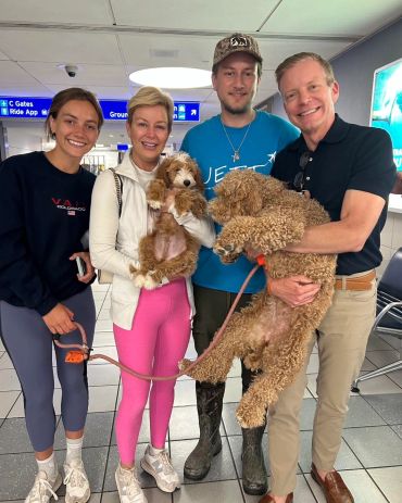Four people at an airport holding two curly-haired dogs, smiling.