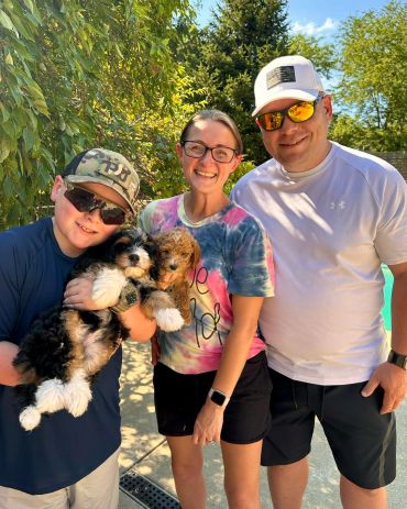 Smiling family outdoors holding two puppies on a sunny day.