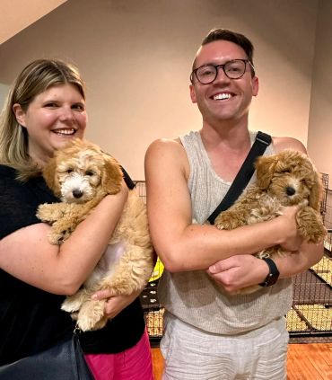 Two smiling people each holding a fluffy puppy indoors.