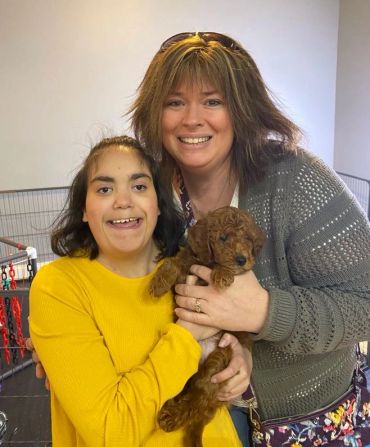 Two smiling women holding a small brown puppy indoors.