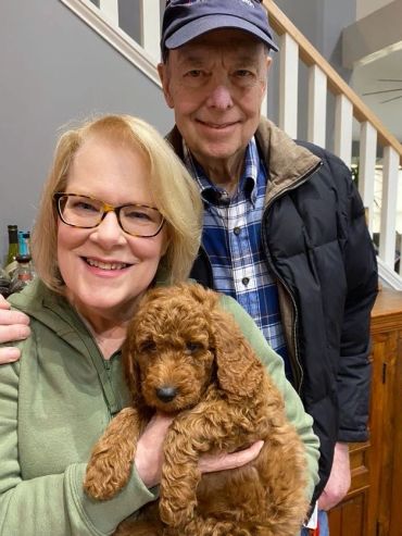 Smiling couple holding a curly-haired brown puppy indoors.