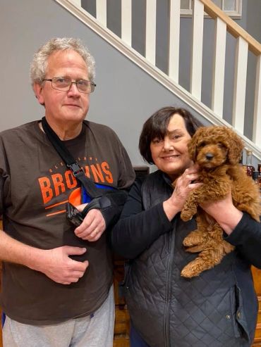 Couple posing indoors with a brown curly-haired puppy, man wearing arm sling.