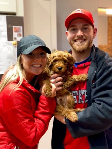 Smiling couple holding an adorable curly-haired puppy indoors.