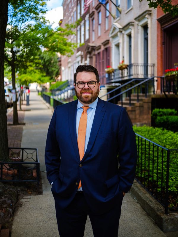 Man in a blue suit and orange tie standing on a city sidewalk.