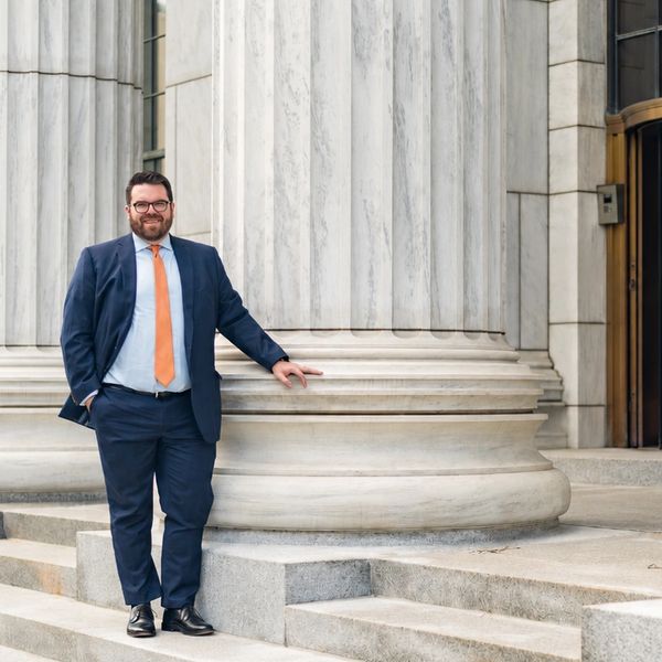 Man in navy suit and orange tie standing by a large marble column.