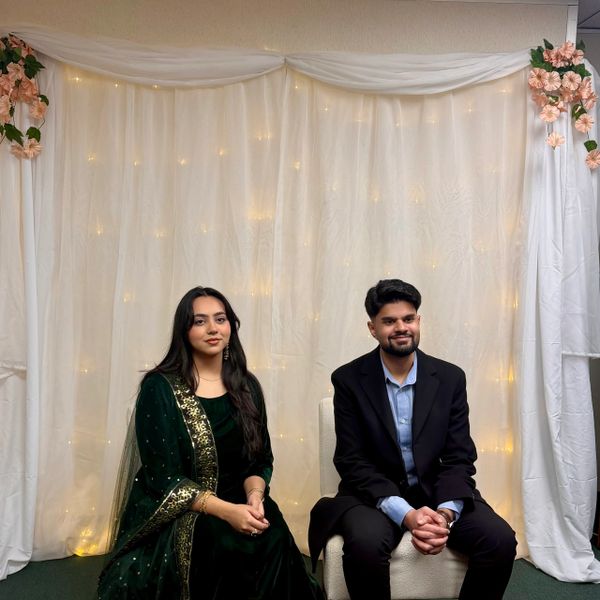 A man and woman dressed formally sitting against a white, softly lit backdrop with floral decorations.