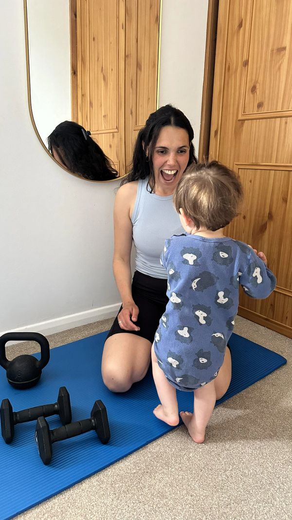 A woman joyfully interacts with a toddler on a yoga mat with weights nearby.
