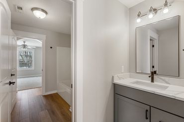 Modern bathroom with gray vanity and light wood flooring leading to a bedroom.