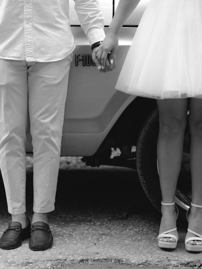 A couple holding hands, dressed elegantly, standing beside a vehicle.