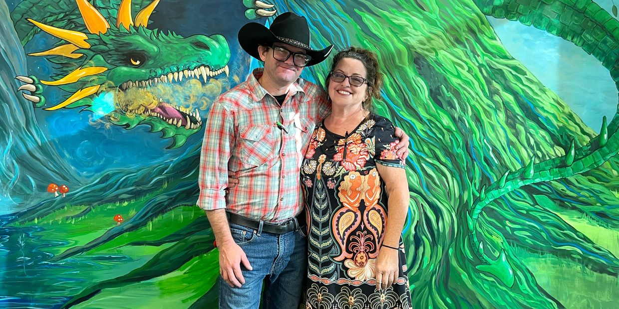 A couple smiling in front of a vibrant dragon mural.