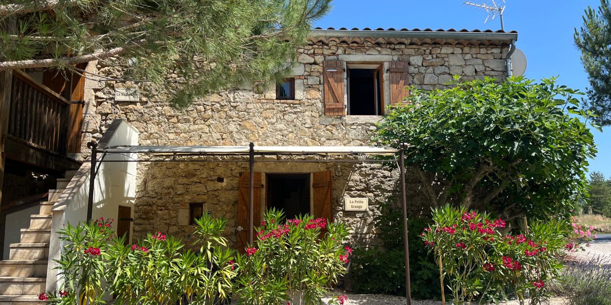 Rustic stone house with wooden shutters surrounded by greenery and flowers under a clear blue sky.