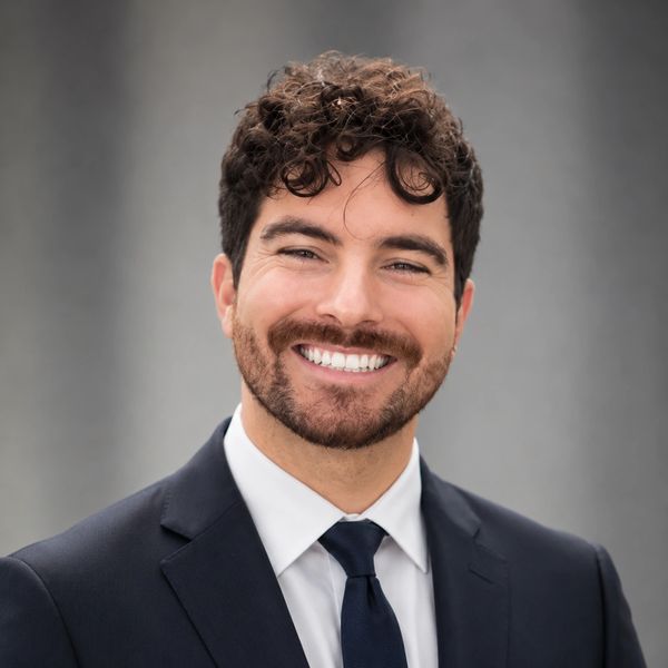 Smiling man in formal suit with curly hair against a gray background.