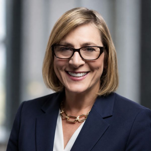 Professional woman with glasses smiling in a navy blazer and gold necklace.