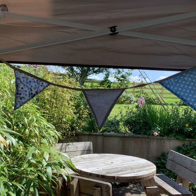 A wooden table & benches, surrounded by green shrubbery under blue sky. Bunting hangs from a parasol