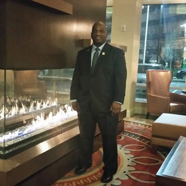 Man in a black suit standing by a modern indoor fireplace.