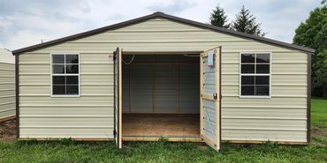 A beige metal storage shed with double doors and windows on a grassy lawn.