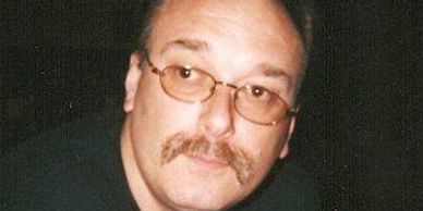Man with glasses and mustache looking at the camera in a dark setting.