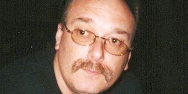 Man with glasses and mustache looking at the camera in a dark setting.