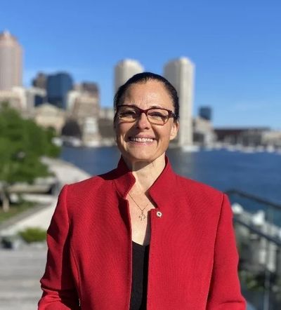 Smiling woman in red jacket by waterfront with city skyline in background.