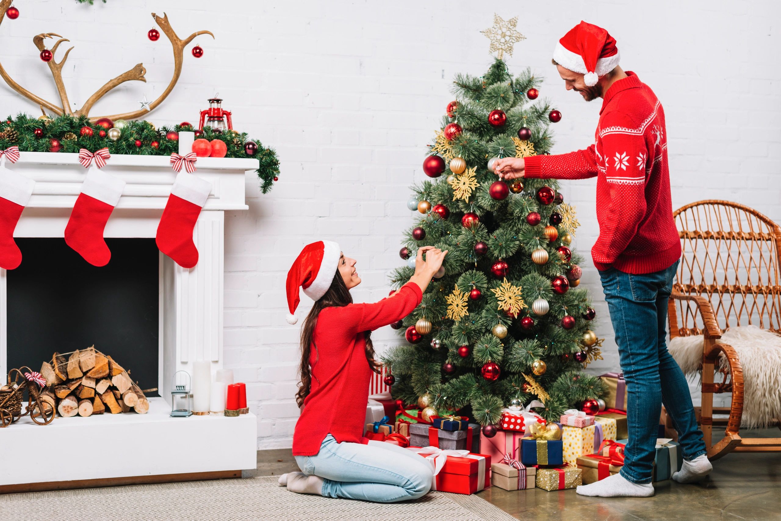 A Couple decorating Christmas Tree wearing Christmas red outfits next to a fireplace.
