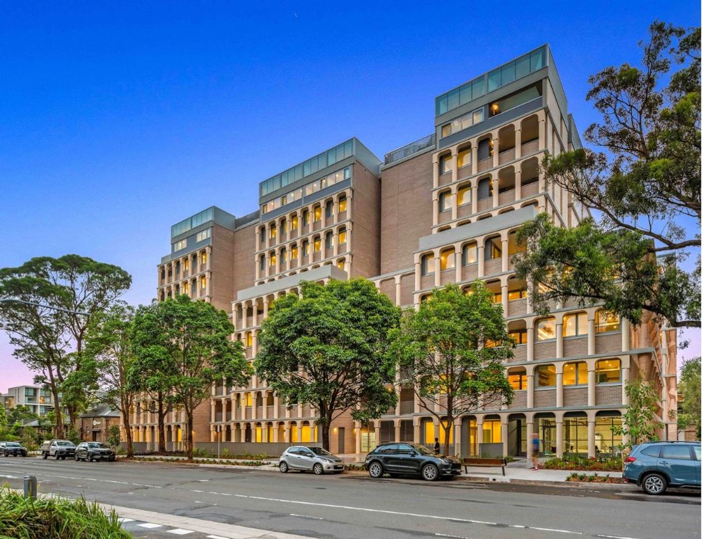 Modern multi-story building with arched windows surrounded by trees and parked cars.