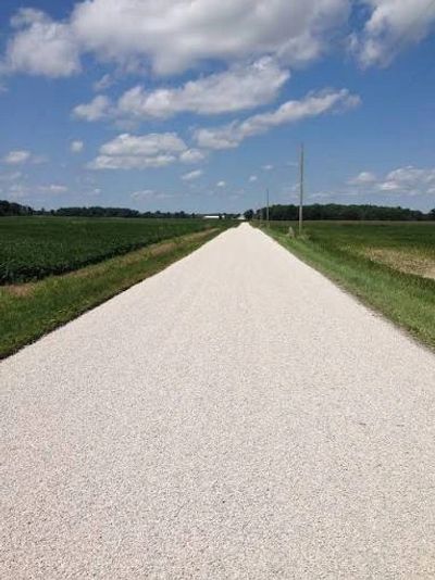 A long, straight gravel road stretches through green fields under a partly cloudy blue sky.