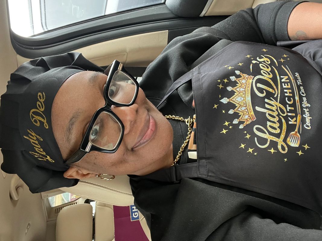 Woman wearing a Lady Dee's Kitchen chef hat and apron smiling in a car.