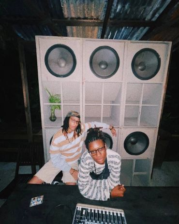 Two women, Steph Z and Marz posing in front of large speakers and a mixer at night.