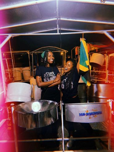 Two smiling women in front of steel drums, one holding a Jamaican flag. Ra'Shain and Irkah in Trinidad 2023