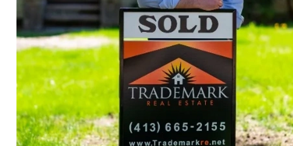Man standing behind a 'Sold' real estate sign on a sunny lawn.