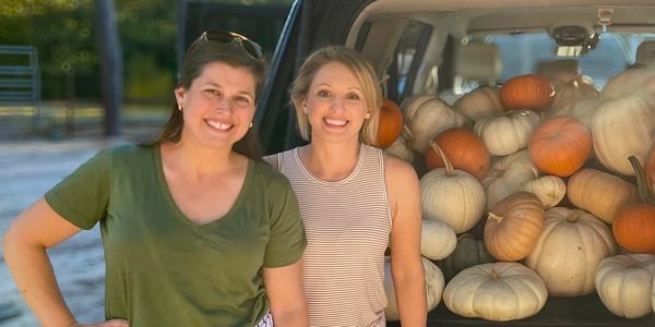 Two women smiling beside a car filled with pumpkins.