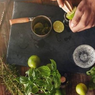 Hands squeezing lime with fresh herbs and glass on a table.