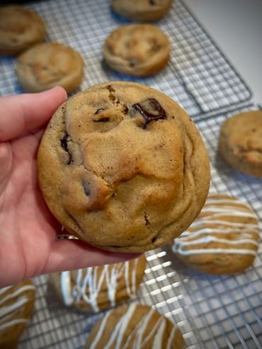 Hand holding a freshly baked chocolate chip cookie with more cookies on cooling racks.