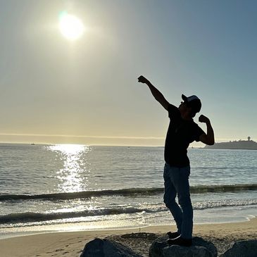 A confidence young man making a body builder pose at the beach, pointing his arm toward the sun.