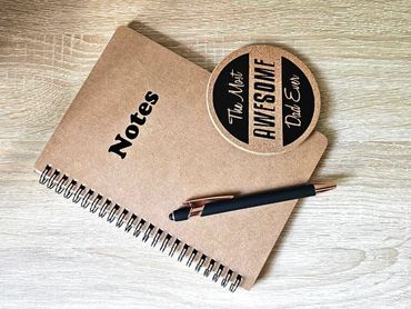 Notebook, pen, and a coaster saying 'The Most Awesome Dad Ever' on a wooden table.