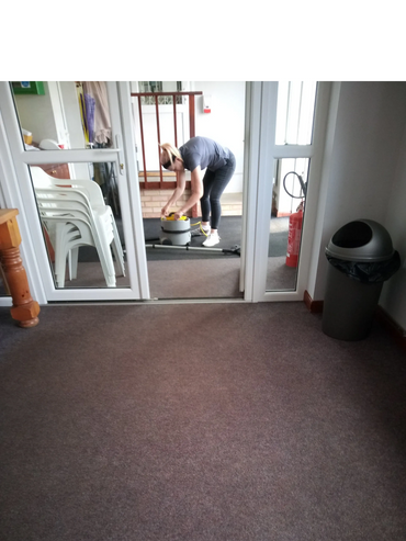 Person setting up a vacuum cleaner in a hallway with stacked chairs and a trash bin.