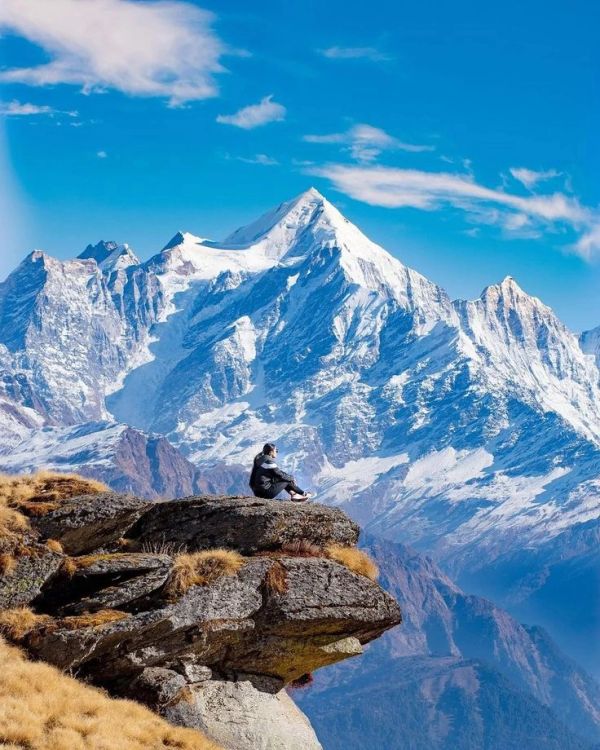 Person sitting on a rocky cliff overlooking snowy mountains.