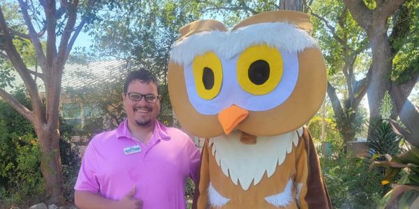 Man in pink shirt posing with a large owl mascot outdoors.